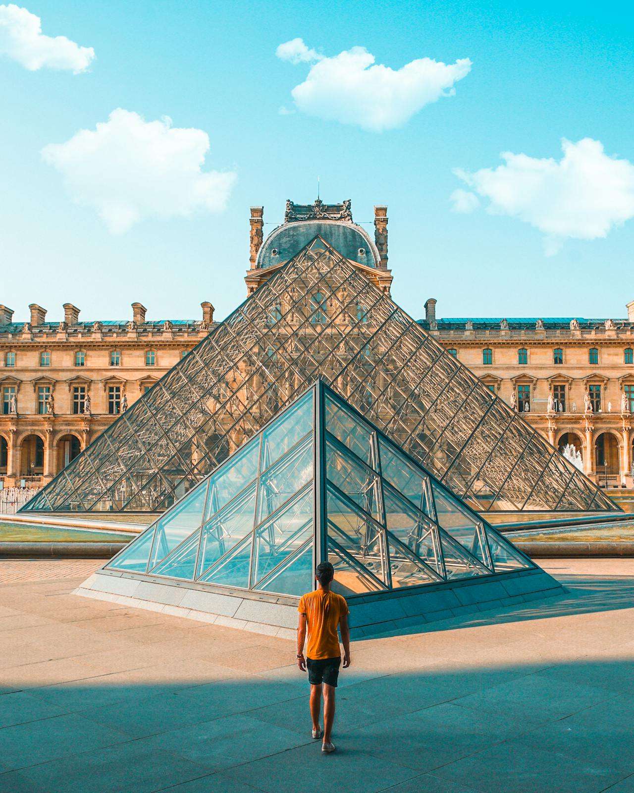 man standing in the middle of the plaza in paris where the lourve is and a glass pyramid around the world top 5 paris france