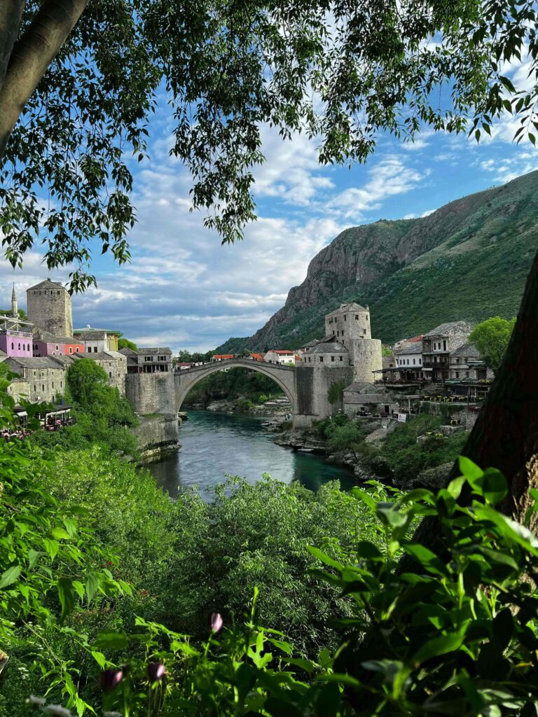 mostar bridge in background with lush green trees and canal under bridge